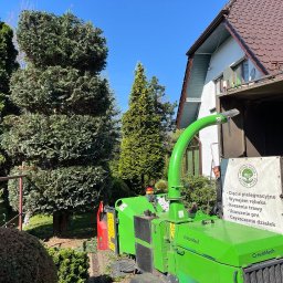 POLSKA FLORA Sp. z O. O. - Zielona rębarka GreenMech przy zaparkowanej przyczepie z napisem 'Wycinka Drzew' na tle wysokich tuj i domu z brązowym dachem.