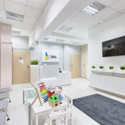 Jasne, minimalistyczne wnętrze poczekalni w placówce medycznej z recepcją, krzesłami dla pacjentów, stolikiem z zabawkami dla dzieci i telewizorem na ścianie.