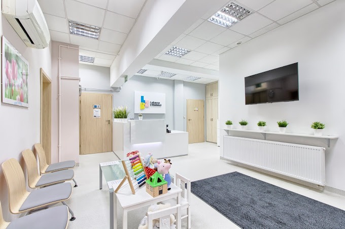 Jasne, minimalistyczne wnętrze poczekalni w placówce medycznej z recepcją, krzesłami dla pacjentów, stolikiem z zabawkami dla dzieci i telewizorem na ścianie.