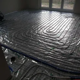 Białe rury ogrzewania podłogowego ułożone w pętle na folii z siatką w nowym budownictwie, widoczne niebieskie rury przy ścianie i okno w tle.