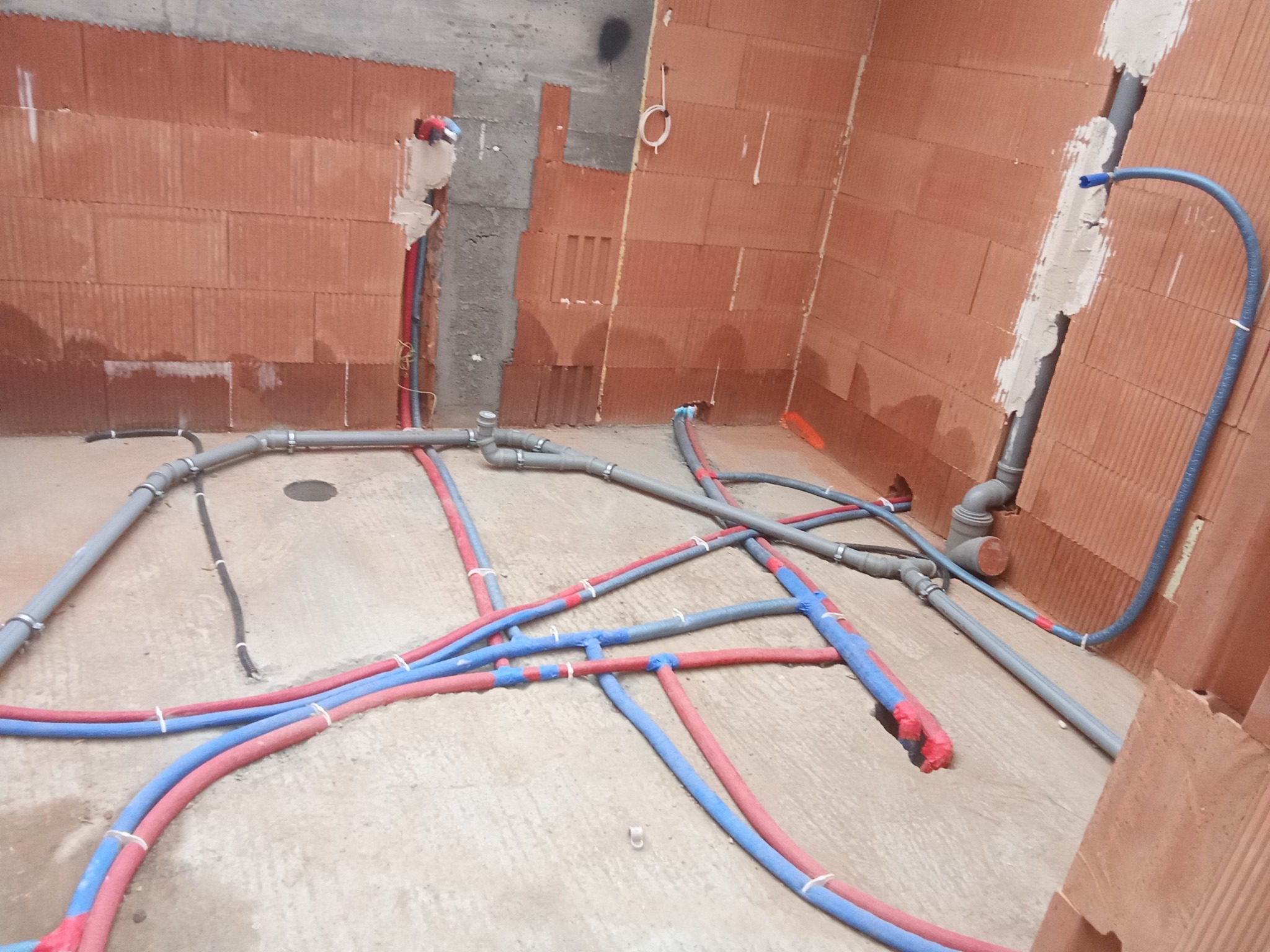 Surowy stan instalacji ogrzewania podłogowego i wodnej w nowym budynku z cegły, rury PEX czerwone i niebieskie połączone tymczasowo białymi opaskami, szare rury kanalizacyjne i wodociągowe