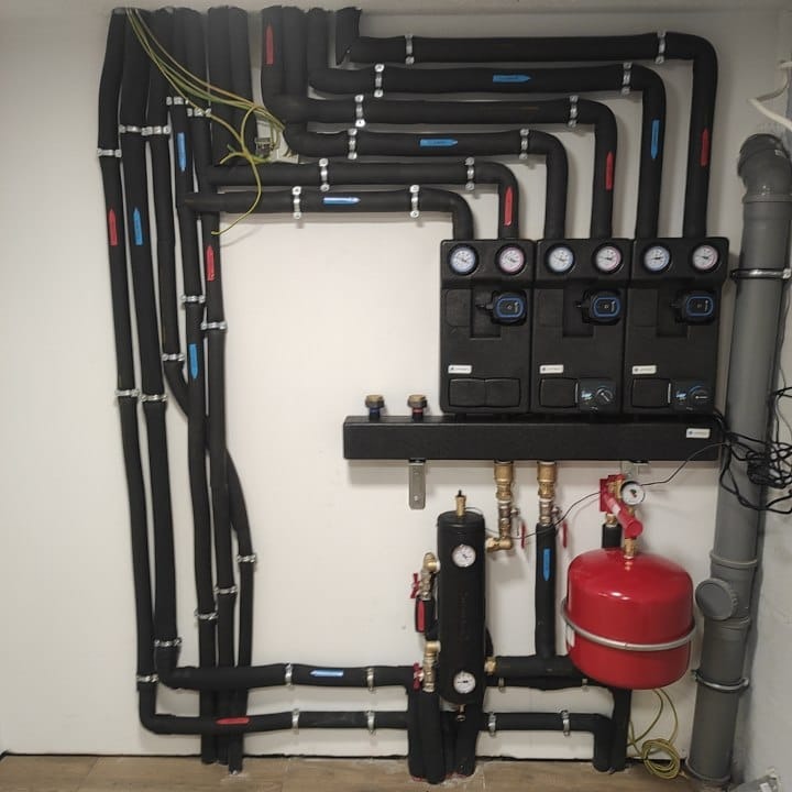 Rozbudowana instalacja grzewcza z czarnymi rurami izolowanymi, rozdzielaczami z manometrami, czerwonym zbiornikiem wyrównawczym i szarą rurą kanalizacyjną w tle.