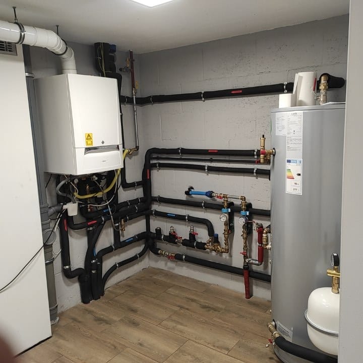 Nowoczesny piec gazowy kondensacyjny z podłączonym zasobnikiem ciepłej wody, widoczne rury w izolacji, zawory i elementy instalacji w kotłowni z drewnopodobną podłogą.