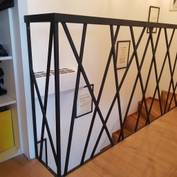 Stylowe schody - Czarna, metalowa balustrada o geometrycznym wzorze krzyżujących się prętów, zabezpieczająca schody z drewnianymi stopniami, widok z góry, obok regał z kaskiem motocyklowym.