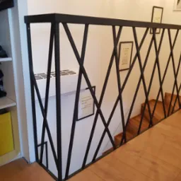 Czarna, metalowa balustrada o geometrycznym wzorze krzyżujących się prętów, zabezpieczająca schody z drewnianymi stopniami, widok z góry, obok regał z kaskiem motocyklowym.