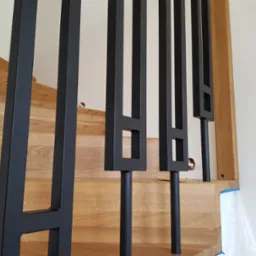 Fragment nowoczesnych schodów z jasnego drewna z czarnymi, metalowymi balustradami o geometrycznym wzorze, widok z bliska na balustrady i schody.