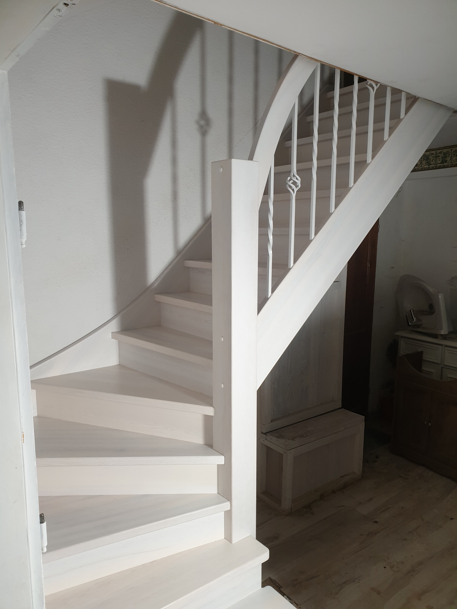 Spiralne, białe schody z balustradą w jasnym wnętrzu, widoczne niedoskonałości malowania i fragmenty starego umeblowania w tle.