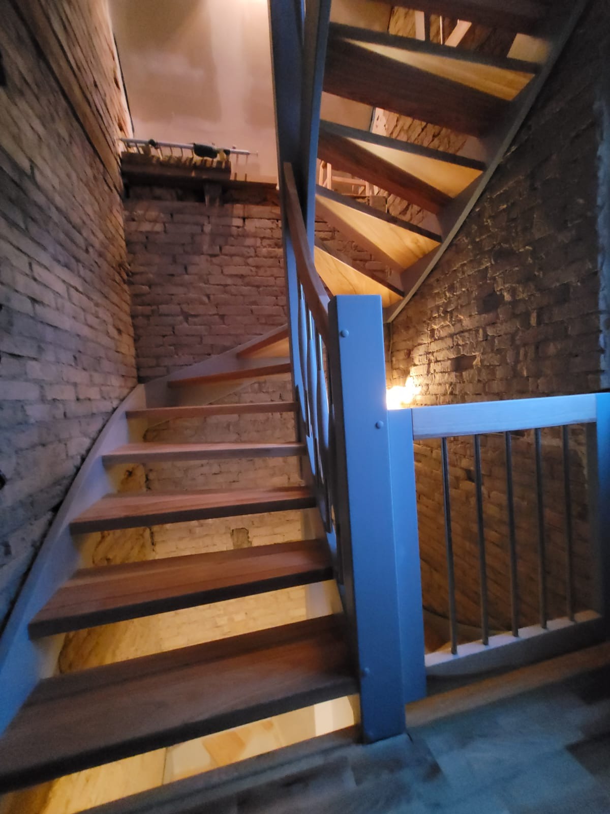 Drewniane schody z szarą balustradą w surowym wnętrzu z ceglanymi ścianami i oświetleniem punktowym pod schodami.