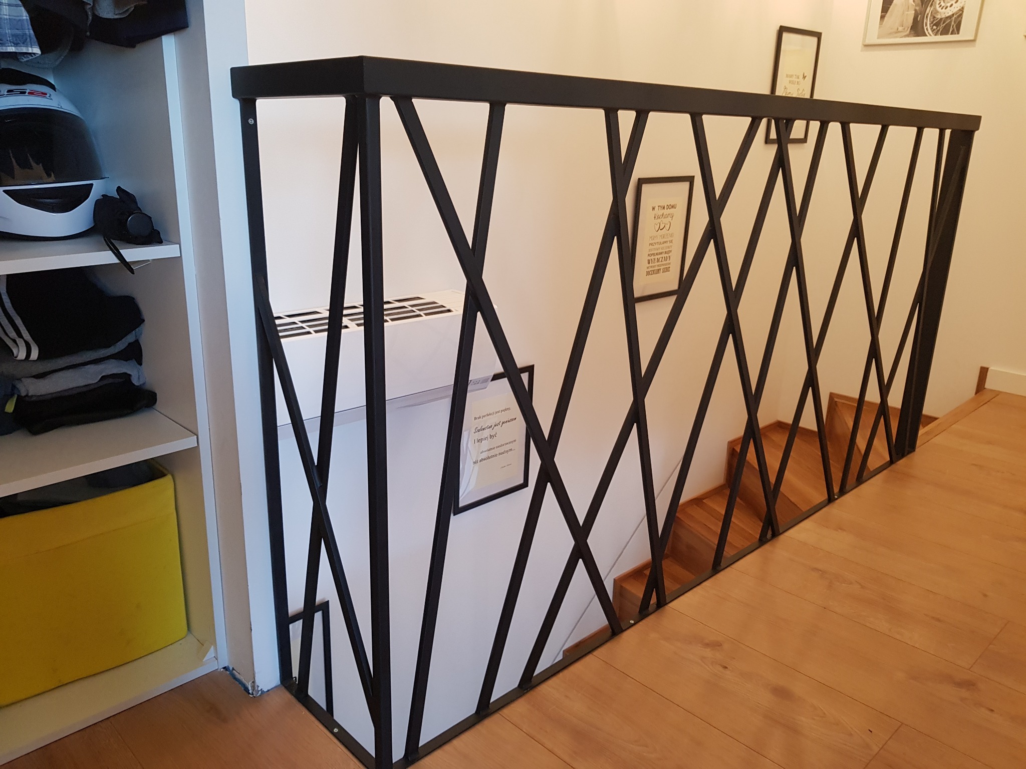 Czarna, metalowa balustrada o geometrycznym wzorze krzyżujących się prętów, zabezpieczająca schody z drewnianymi stopniami, widok z góry, obok regał z kaskiem motocyklowym.