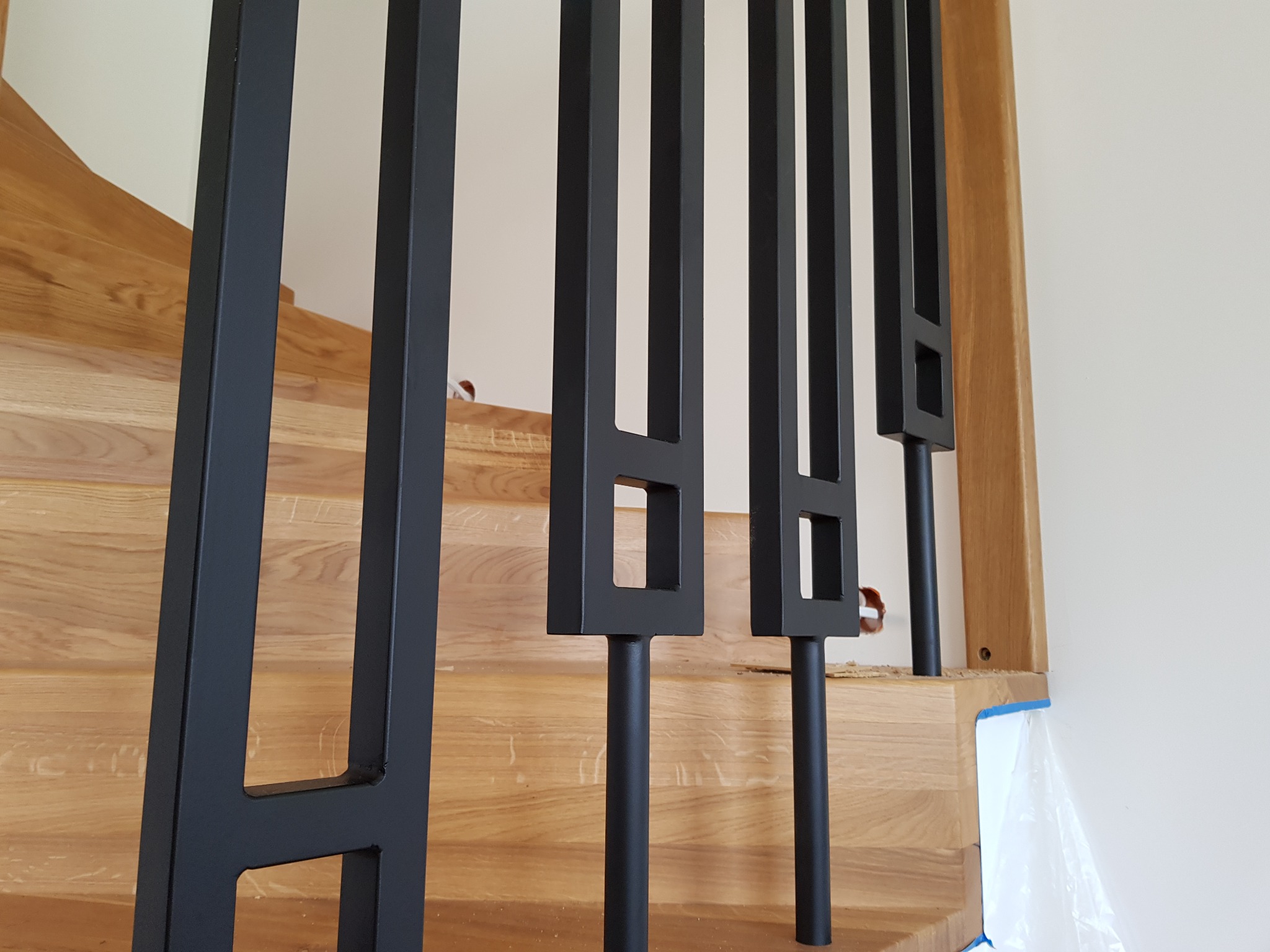 Fragment nowoczesnych schodów z jasnego drewna z czarnymi, metalowymi balustradami o geometrycznym wzorze, widok z bliska na balustrady i schody.