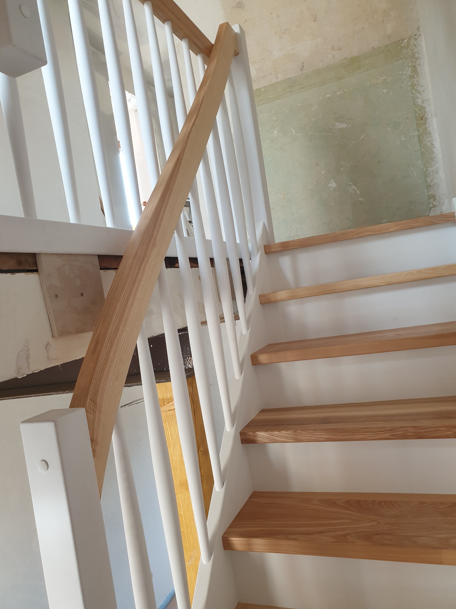 Nowoczesne schody z drewnianymi stopniami i białymi balustradami z okrągłymi szczeblami, widoczne ślady po remoncie na ścianie w tle.