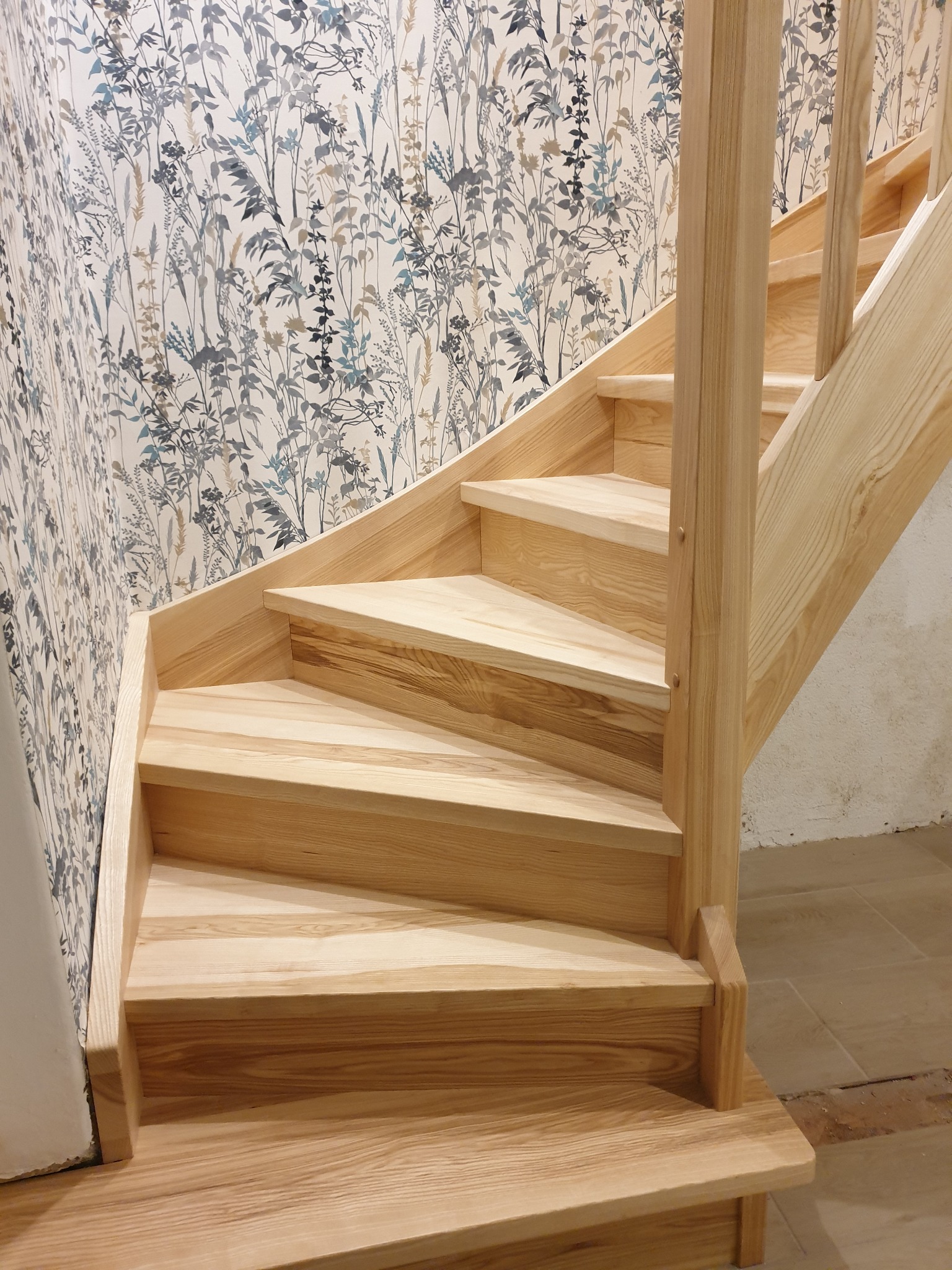 Jasne, drewniane schody z poręczą, widoczne z dołu, z ozdobną tapetą w tle.