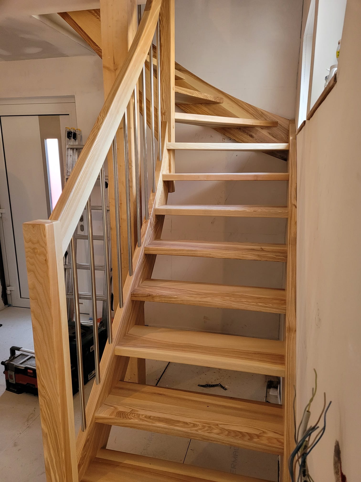 Jasne, drewniane schody z metalowymi balustradami, widok z dołu, w tle drabina i narzędzia, ściany w trakcie wykończenia.