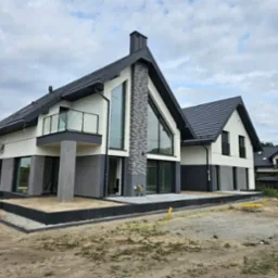 Nowoczesny dom jednorodzinny z elewacją w kolorach bieli i szarości, z dużymi oknami i balkonem z szklaną balustradą, widoczny na etapie wykończenia terenu wokół budynku.