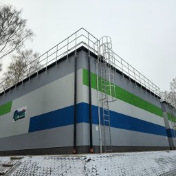 REMONTEAM ELEWACJE SP. Z.O.O. - Duży budynek przemysłowy z elewacją w pasy szare, białe, zielone i niebieskie, z logo firmy na ścianie, drabina na elewacji, otoczony drzewami i pokryty śniegiem.