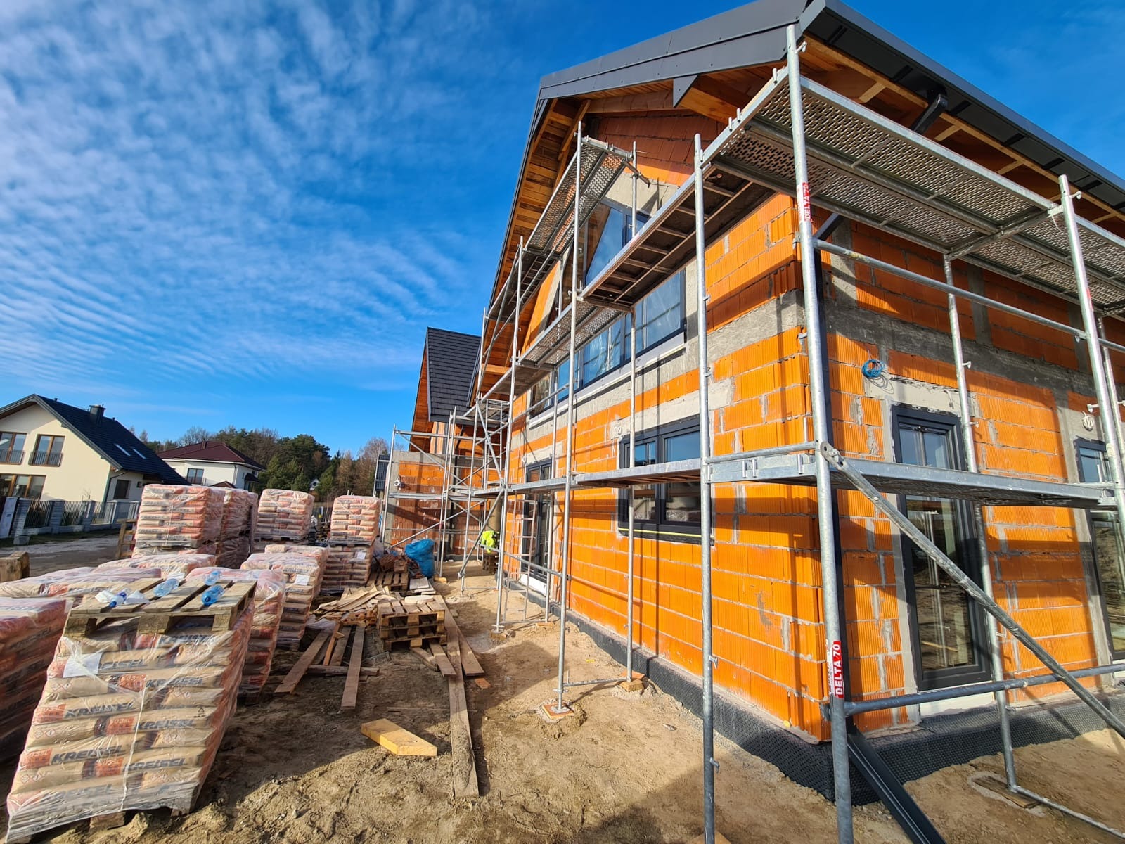 Dom jednorodzinny w trakcie budowy, widoczne pomarańczowe pustaki ceramiczne, szare spoiny, czarne okna, rusztowanie, palety z zaprawą murarską, błękitne niebo z chmurami.