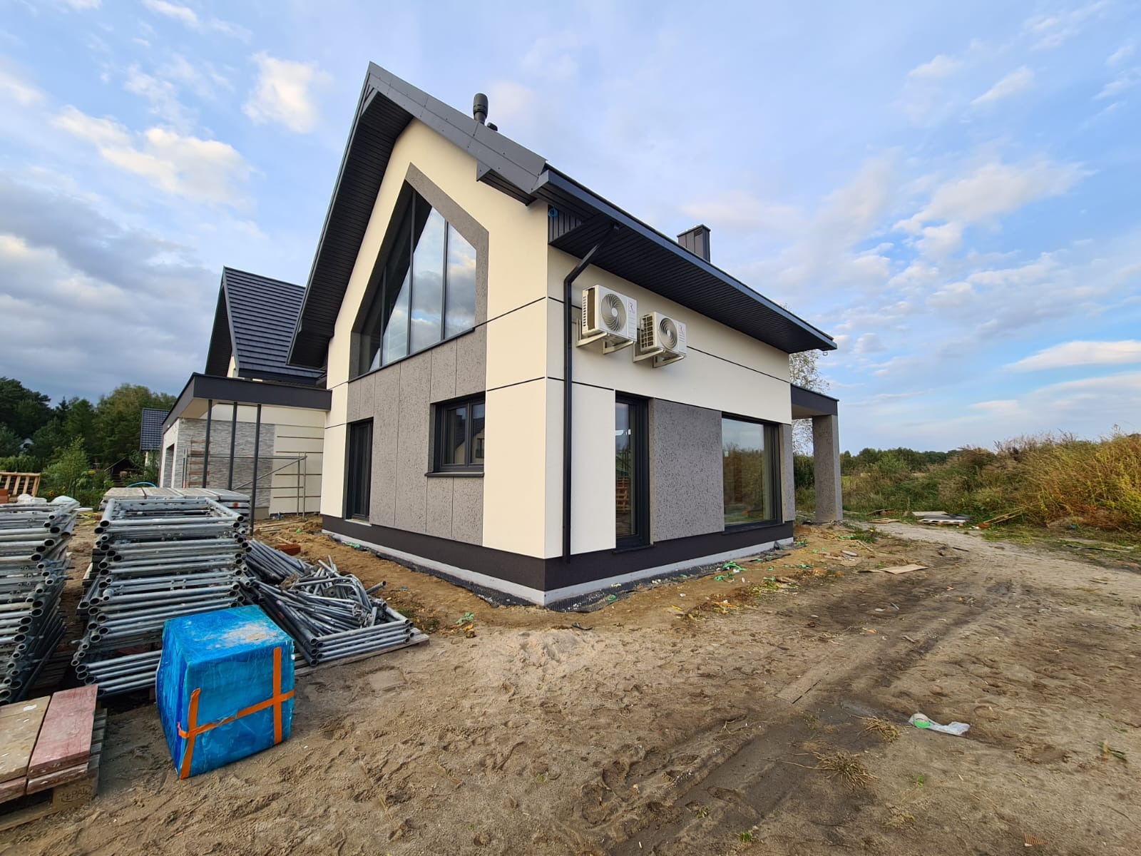 Nowoczesny dom z elewacją w odcieniach szarości i beżu, z widocznymi jednostkami klimatyzacji na ścianie oraz stertą rusztowań i materiałów budowlanych na pierwszym planie.