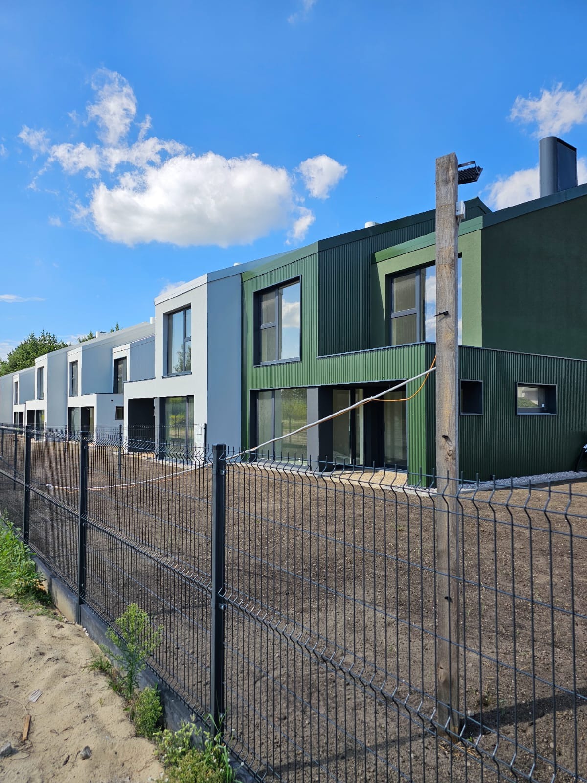 Szereg nowo wybudowanych domów bliźniaczych z elewacją w odcieniach szarości i zieleni, widocznych za metalowym ogrodzeniem panelowym. Teren przed domami przygotowany pod trawnik. Słup...