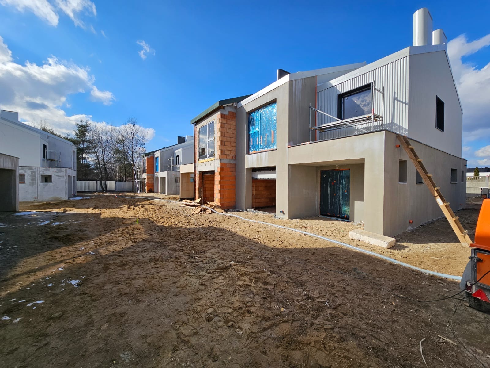 Rząd szeregowych domów w trakcie budowy, z widocznymi elementami elewacji w różnym stadium zaawansowania, od surowej cegły po otynkowane i częściowo pomalowane ściany, zabezpieczone okna folią...