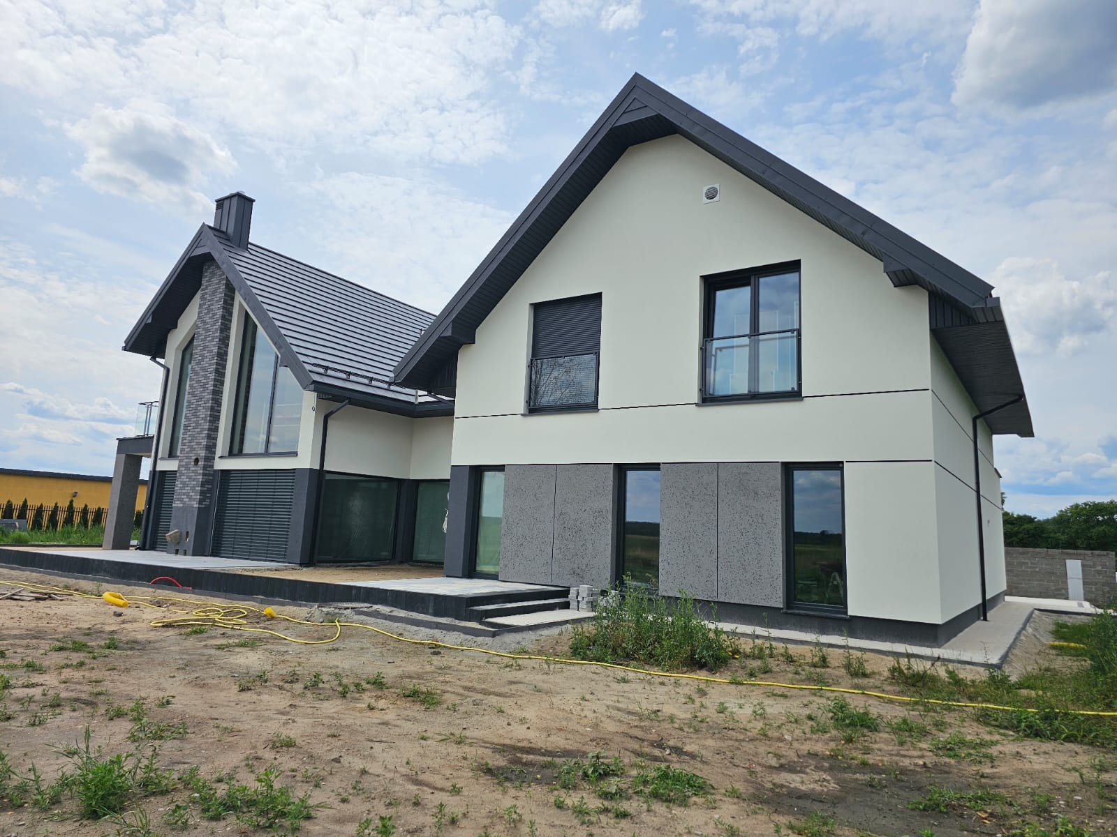 Nowoczesny dom jednorodzinny z jasną elewacją, ciemnymi elementami wykończeniowymi i fragmentami elewacji wykończonymi szarą strukturą, widoczne okna z czarnymi ramami i fragment tarasu z kostki...