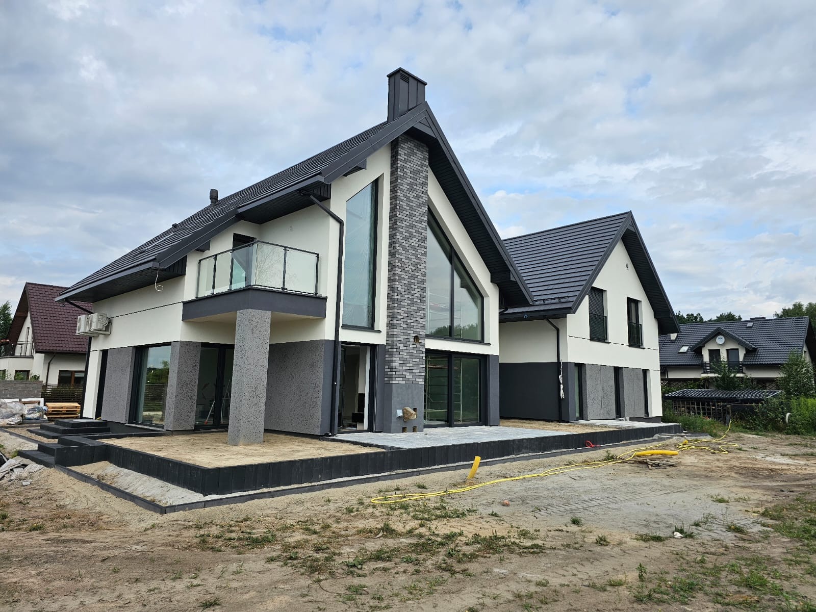 Nowoczesny dom jednorodzinny z elewacją w kolorach bieli i szarości, z dużymi oknami i balkonem z szklaną balustradą, widoczny na etapie wykończenia terenu wokół budynku.