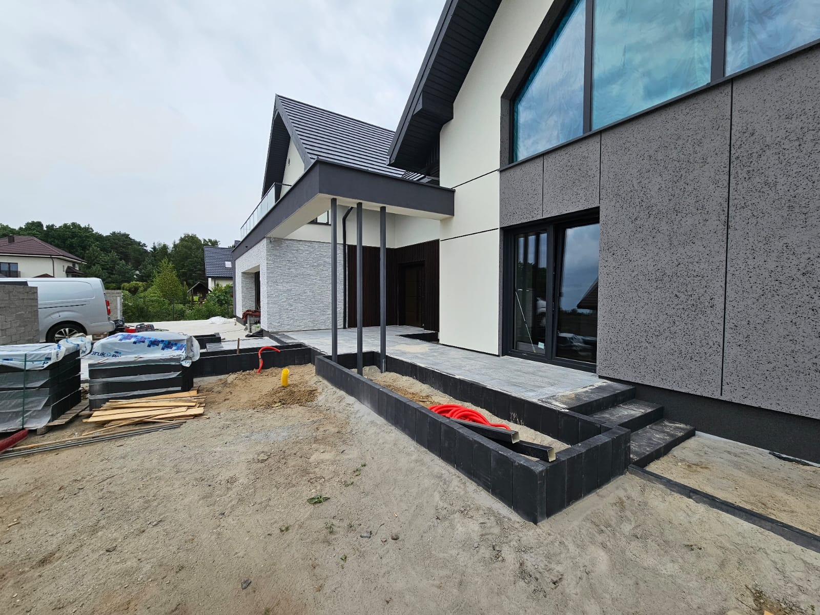 Nowoczesny dom jednorodzinny z elewacją z szarego tynku strukturalnego i białych paneli, widoczne prace przygotowawcze do zagospodarowania terenu przed domem, w tle zaparkowany biały bus.