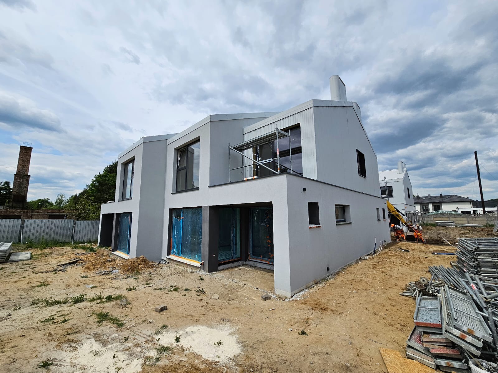 Nowoczesny dom jednorodzinny z szarą elewacją, okna zabezpieczone folią, rusztowania w tle, teren wokół domu w trakcie prac wykończeniowych.