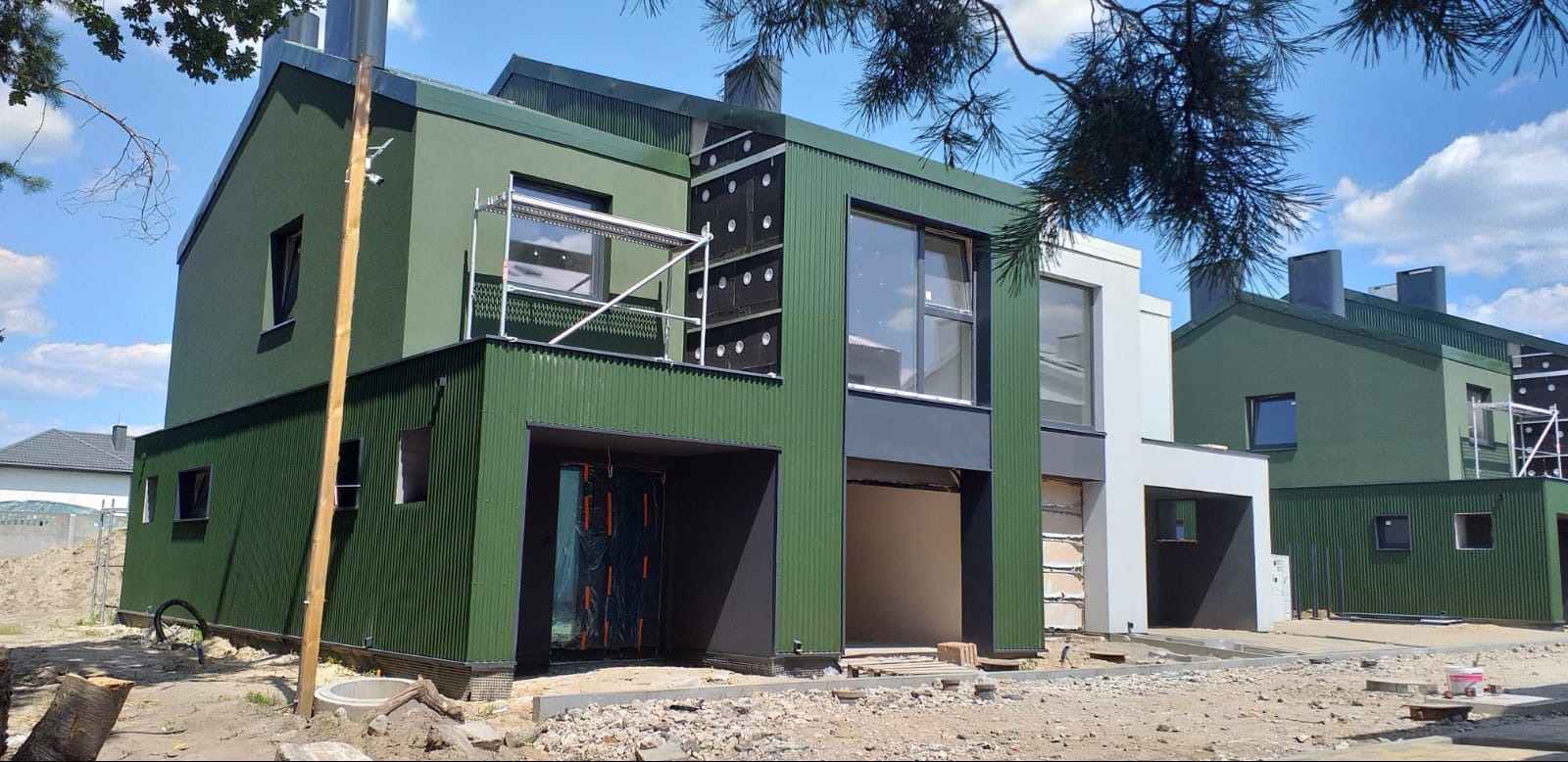 Nowoczesny dom w trakcie wykańczania elewacji: zielona blacha falista na parterze, szara gładka elewacja na piętrze, widoczne rusztowanie i ocieplenie w górnej części, teren budowy wokół.