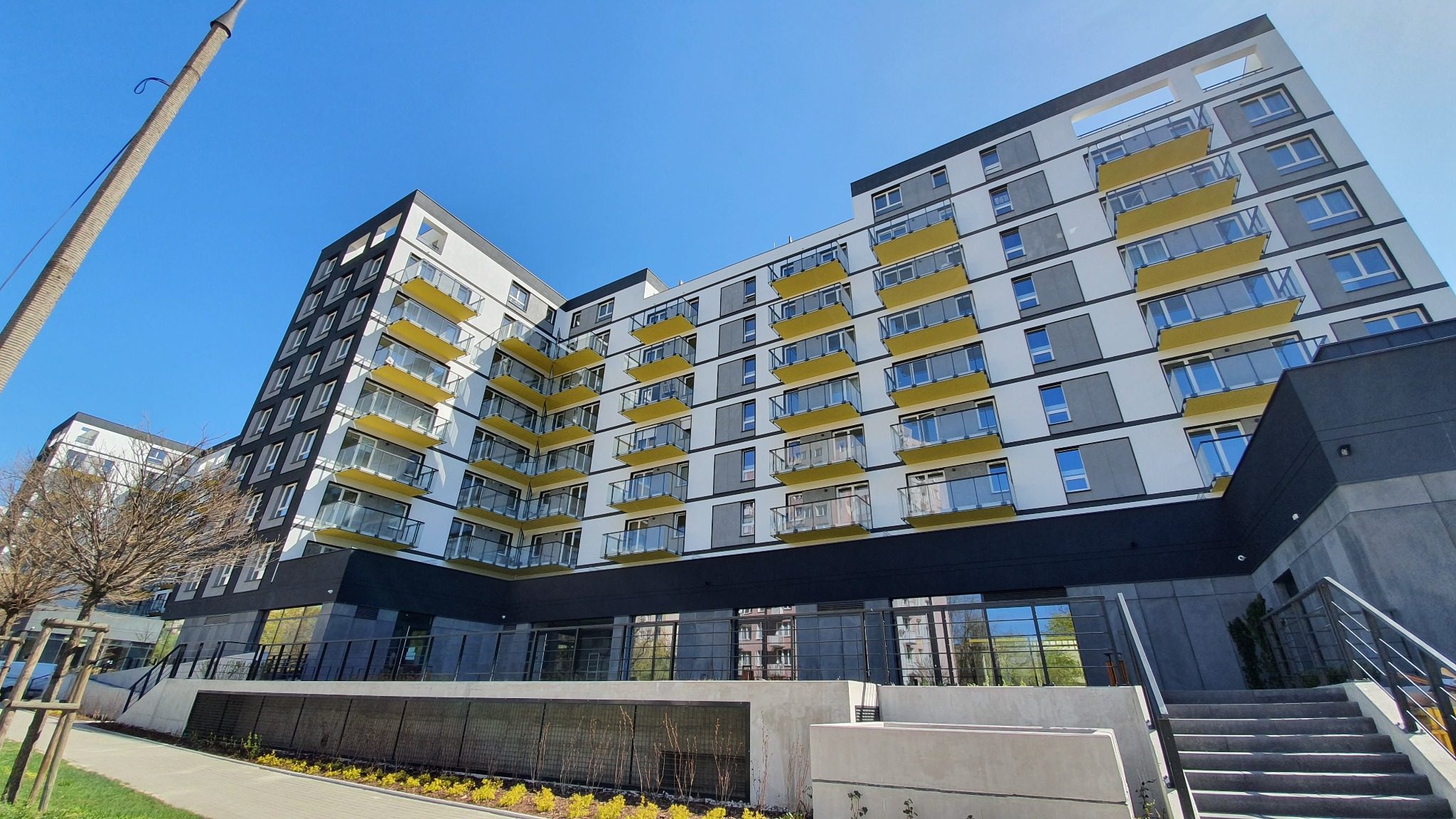 Nowoczesny budynek wielorodzinny w Zielonce z białoszarą elewacją i żółtymi balkonami, sfotografowany pod kątem od dołu w słoneczny dzień.
