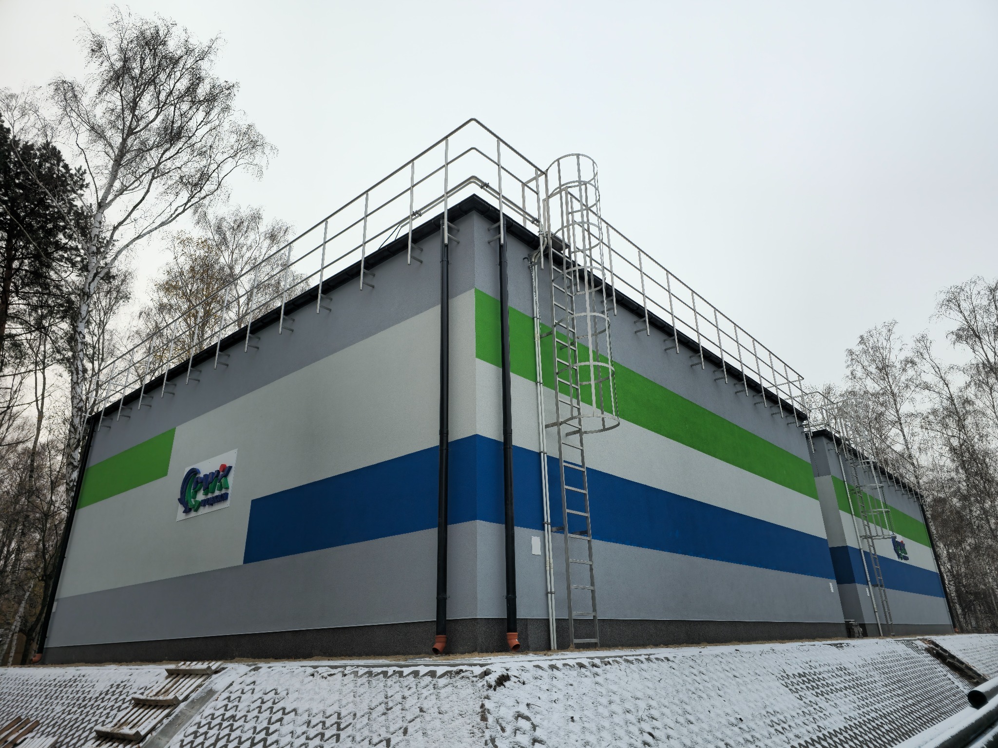 Duży budynek przemysłowy z elewacją w pasy szare, białe, zielone i niebieskie, z logo firmy na ścianie, drabina na elewacji, otoczony drzewami i pokryty śniegiem.