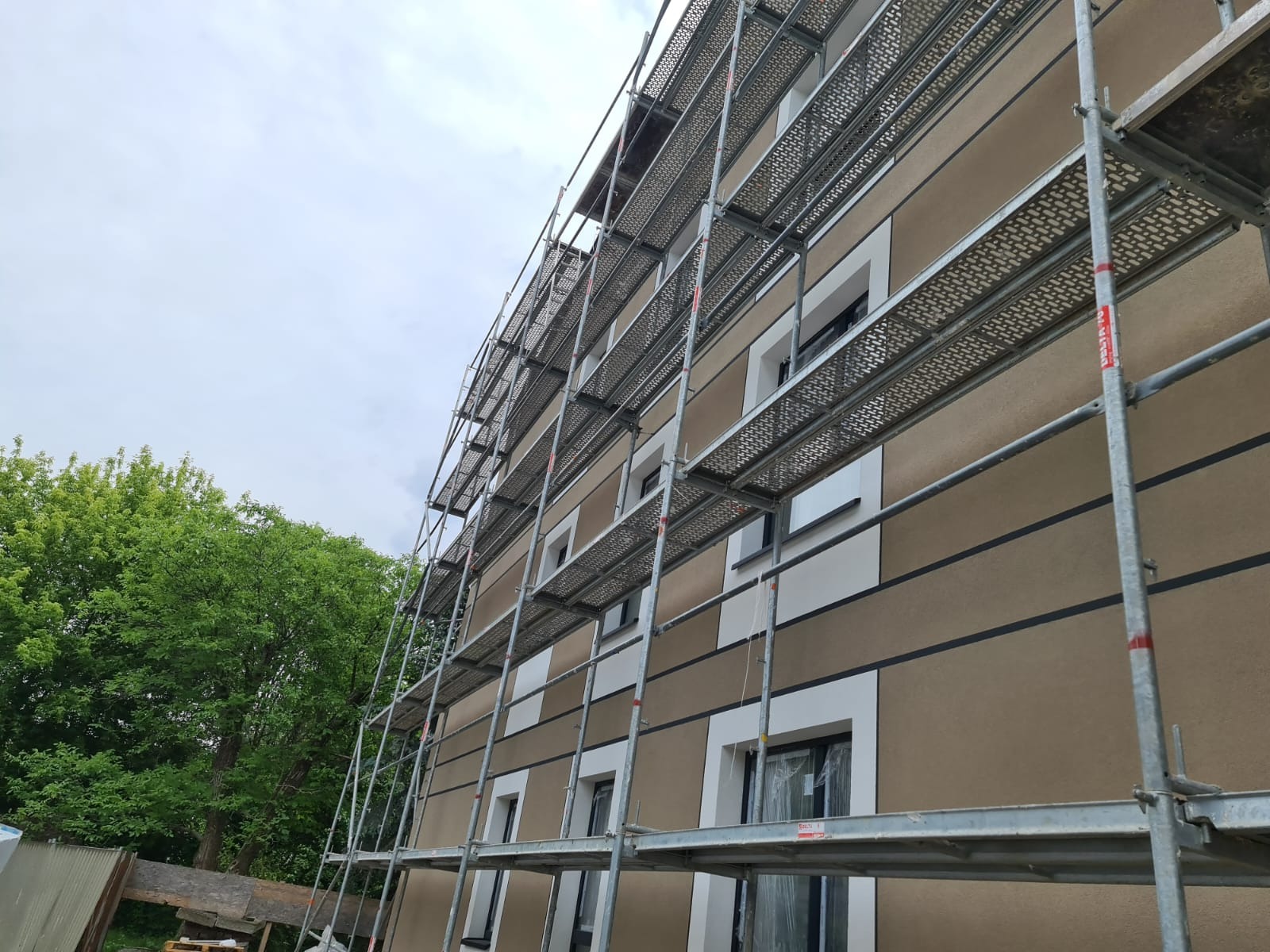 Budynek wielorodzinny w trakcie prac elewacyjnych, widoczne rusztowania, brązowa elewacja z białymi obramowaniami okien i ciemnymi pasami, pochmurne niebo.