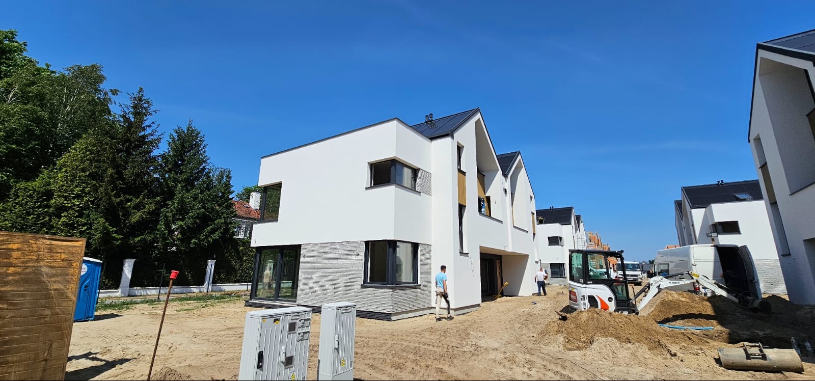 Nowoczesne domy w zabudowie szeregowej z białymi elewacjami i grafitowymi dachami, teren wokół w trakcie prac wykończeniowych, widoczna mini koparka.