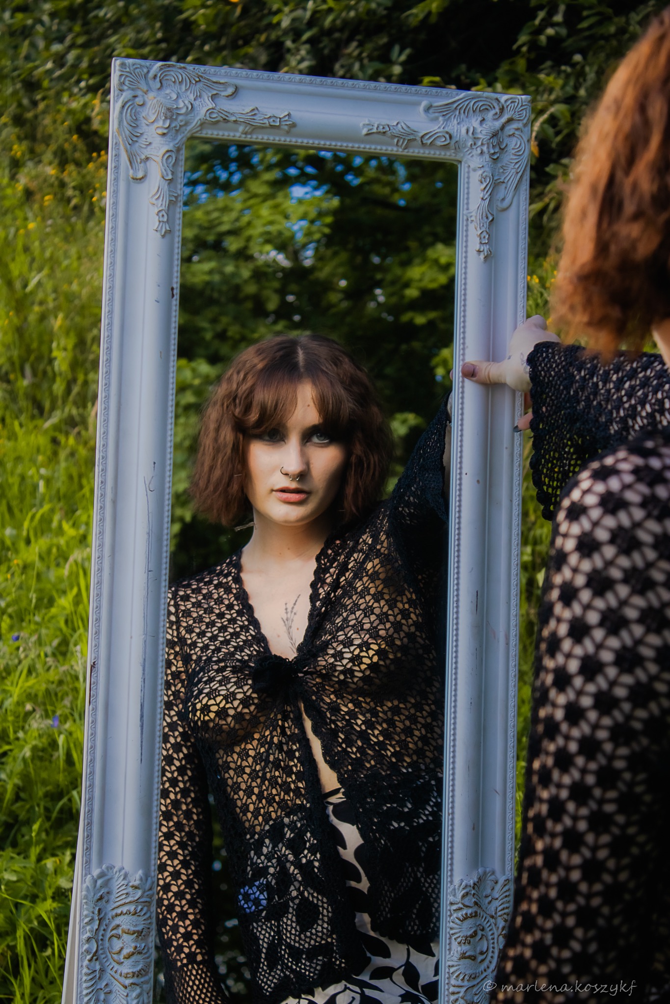 Portret kobiety z piercingiem w nosie, odbijającej się w ozdobnym lustrze na tle zieleni. Kobieta ubrana w ażurowy sweter. Ujęcie w plenerze, naturalne światło.