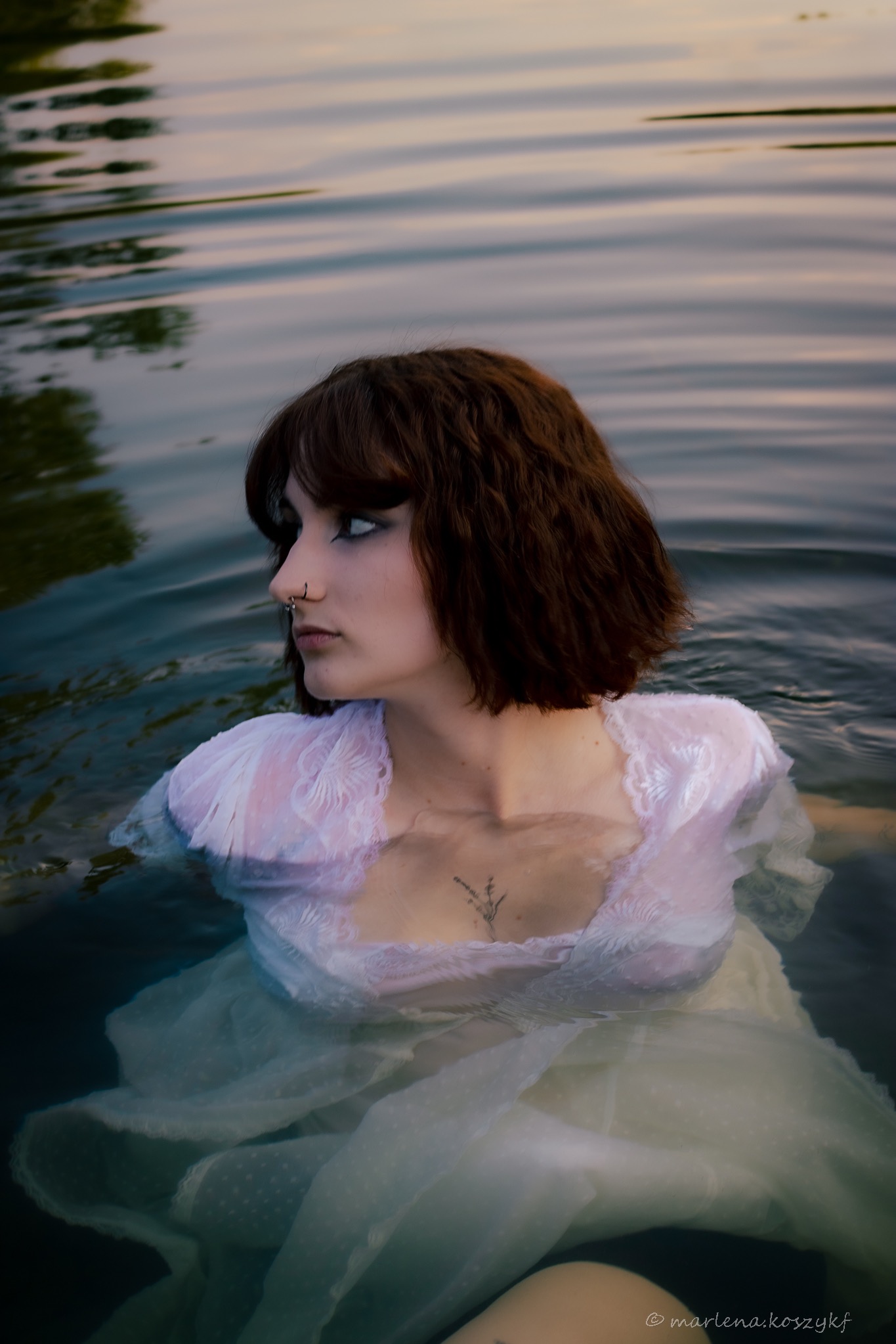 Młoda kobieta zanurzona w wodzie, ubrana w białą, koronkową sukienkę, z widocznym tatuażem na dekolcie i kolczykiem w nosie, spogląda w lewo na tle falującej wody.