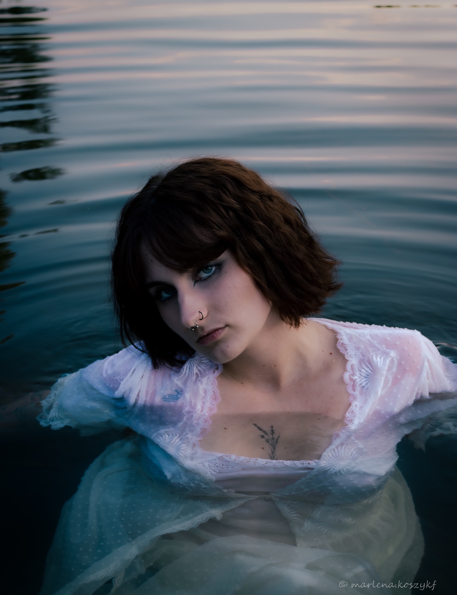 Portret kobiety zanurzonej w wodzie w koronkowej sukience, z piercingiem nosa i tatuażem na dekolcie, patrzącej w obiektyw z melancholijnym wyrazem.