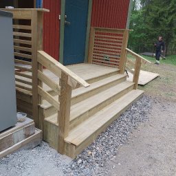 Patryk Torzewski Prestigio - Nowe, drewniane schody wejściowe z balustradą, prowadzące do czerwonego domu z niebieskimi drzwiami, widok z boku, na żwirowym podłożu, w tle mężczyzna z telefonem.