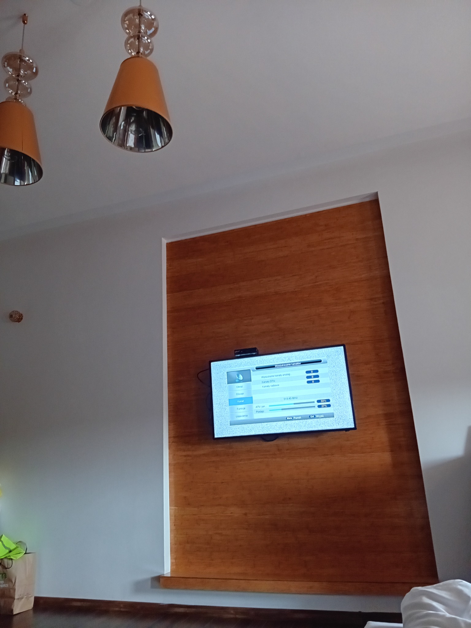 Nowoczesny salon z drewnianą ścianą akcentującą, na której wisi telewizor, oświetlony przez dwie lampy wiszące z elementami szkła i metalu.