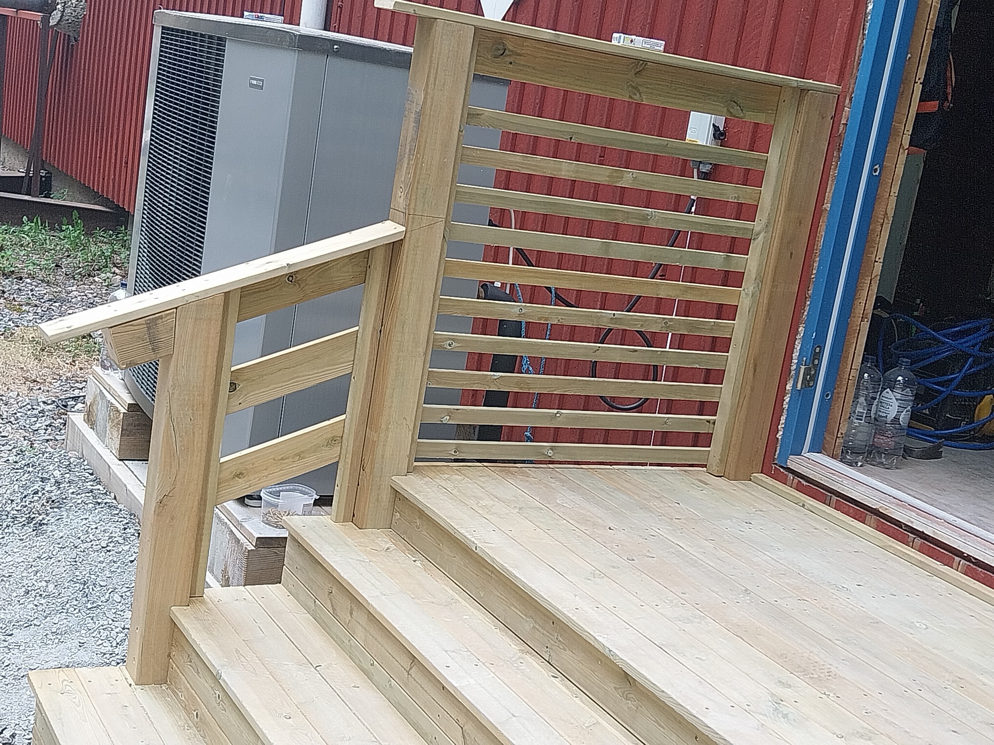 Nowo wybudowany drewniany taras z balustradą, widoczne schody i klimatyzator na zewnątrz budynku z czerwoną elewacją.