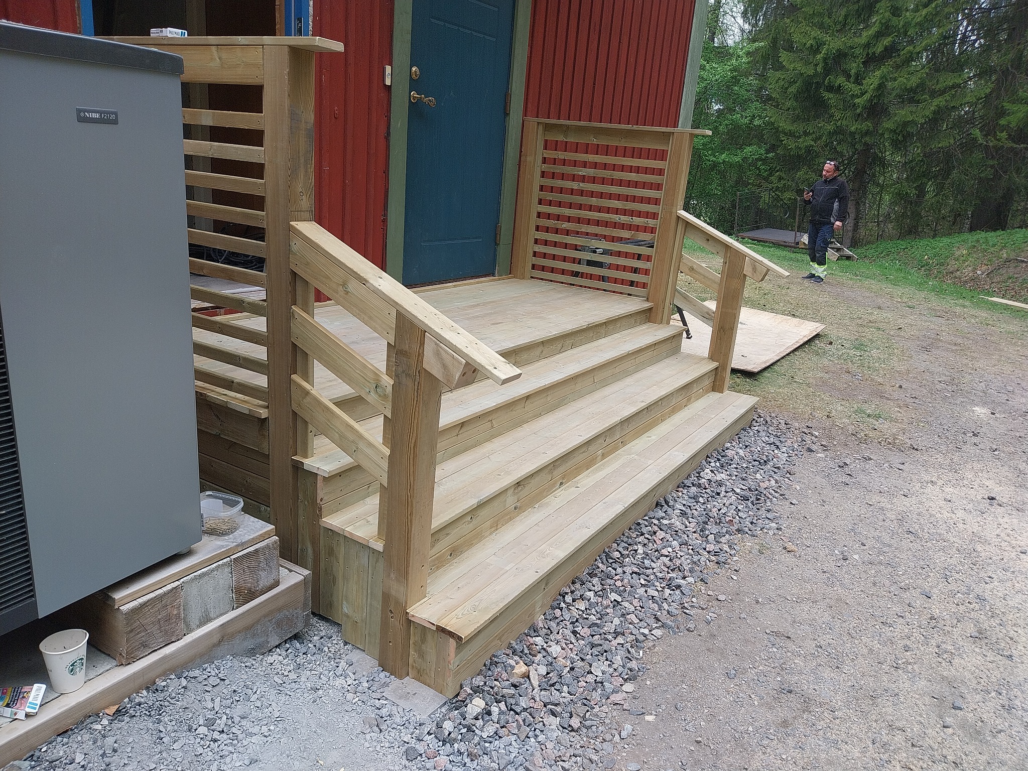 Nowe, drewniane schody wejściowe z balustradą, prowadzące do czerwonego domu z niebieskimi drzwiami, widok z boku, na żwirowym podłożu, w tle mężczyzna z telefonem.