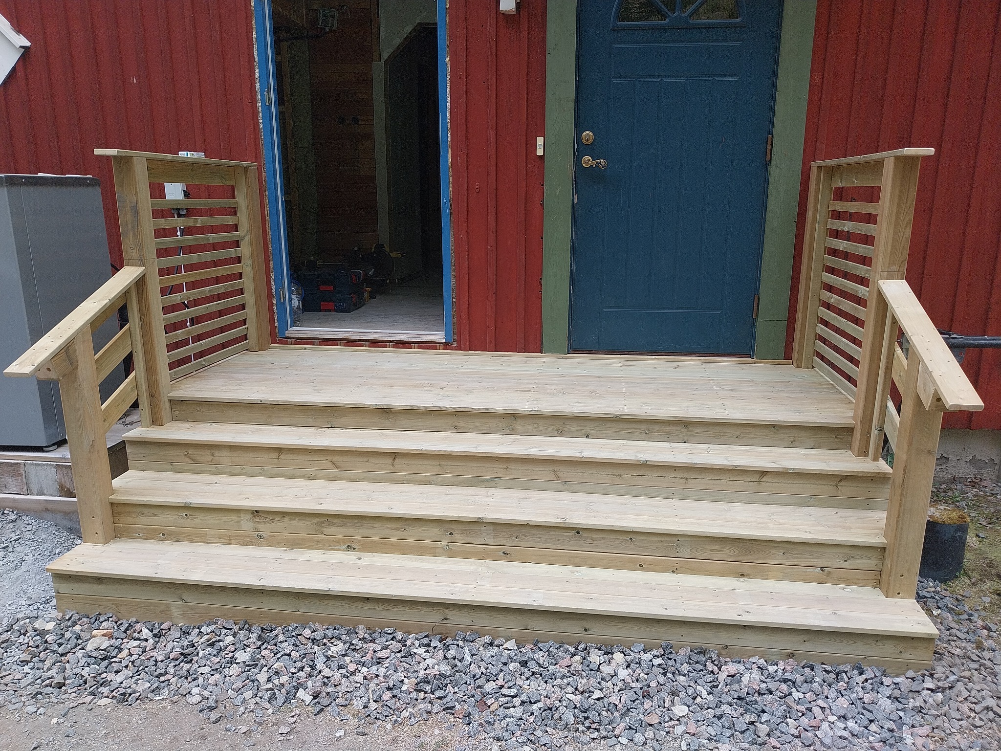 Nowe, drewniane schody wejściowe z balustradami prowadzące do budynku z czerwoną elewacją i niebieskimi drzwiami.