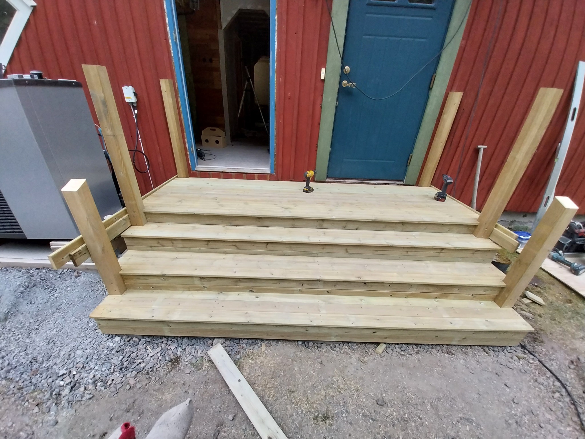 Drewniane schody wejściowe w trakcie budowy, z widocznymi narzędziami, takimi jak wiertarka akumulatorowa, oraz elementami konstrukcyjnymi, na tle czerwonej ściany budynku i niebieskich drzwi.