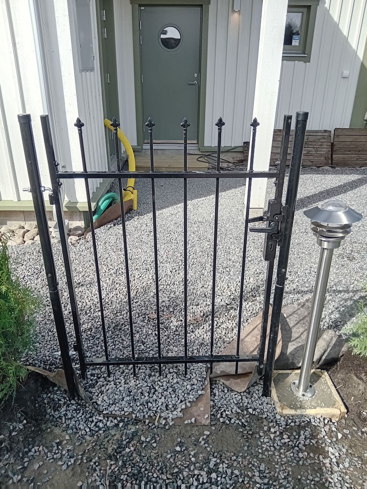 Nowo zainstalowana czarna metalowa furtka wejściowa z ozdobnymi grotami, na żwirowym podłożu przed domem z szarą elewacją i zielonymi drzwiami, widoczny fragment lampy ogrodowej