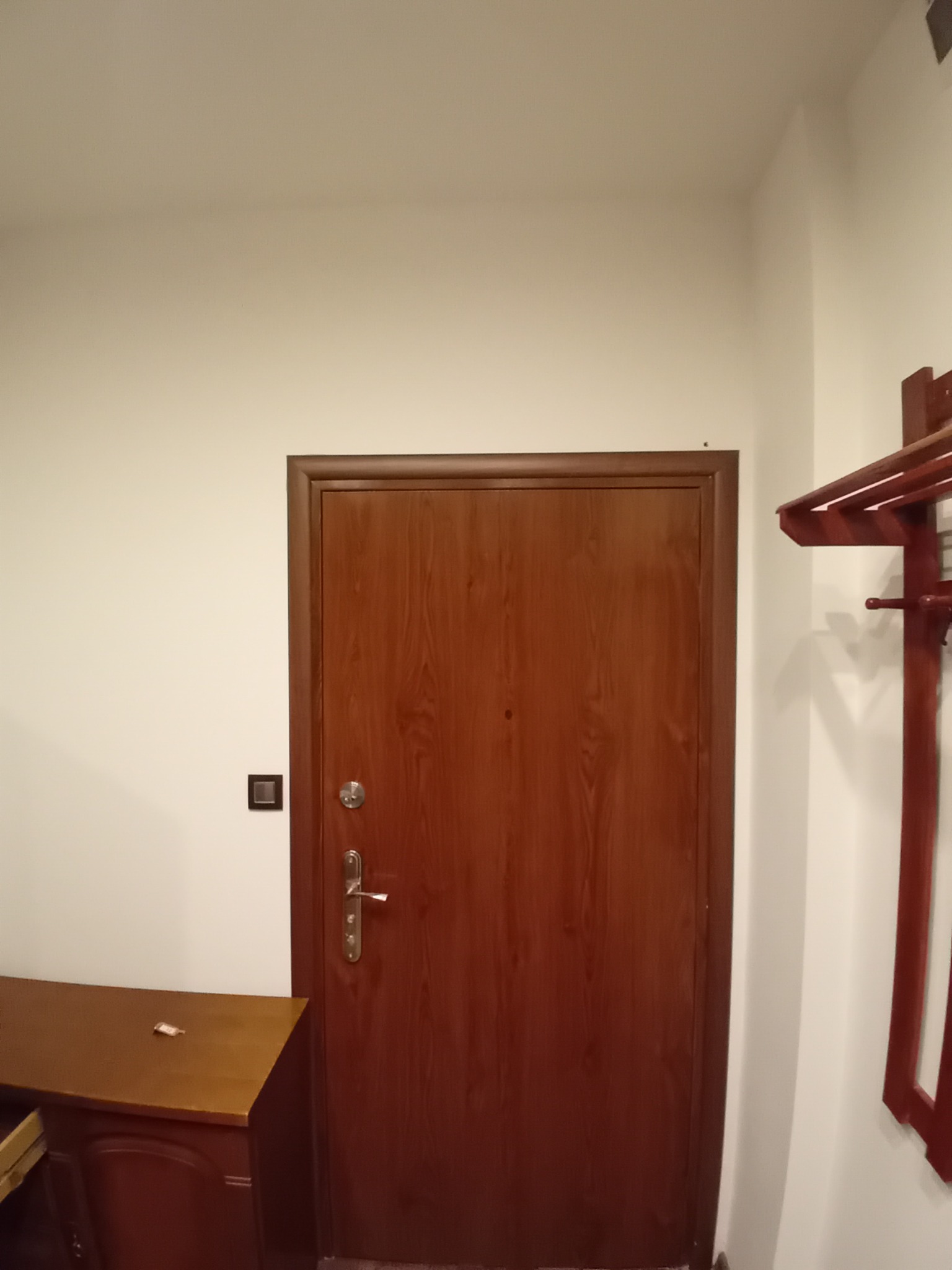 Brązowe drzwi wewnętrzne z ościeżnicą w jasnym korytarzu, widoczny fragment wieszaka na ubrania i drewnianej komody.