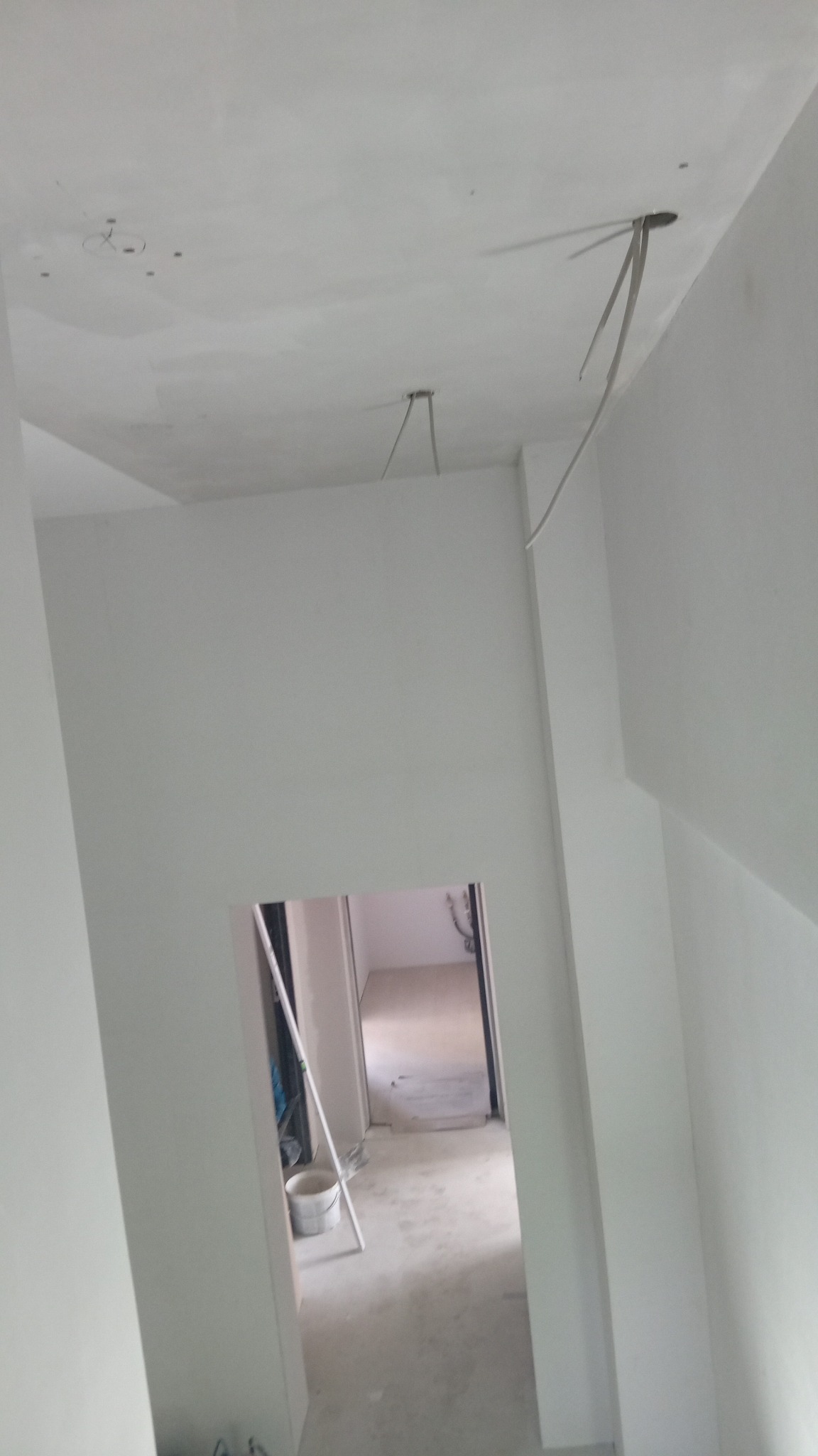 Wnętrze w trakcie remontu: białe ściany po szpachlowaniu, widoczne kable wystające z sufitu, przygotowane do montażu oświetlenia, w tle widoczne pomieszczenie z wiadrem i drabina.