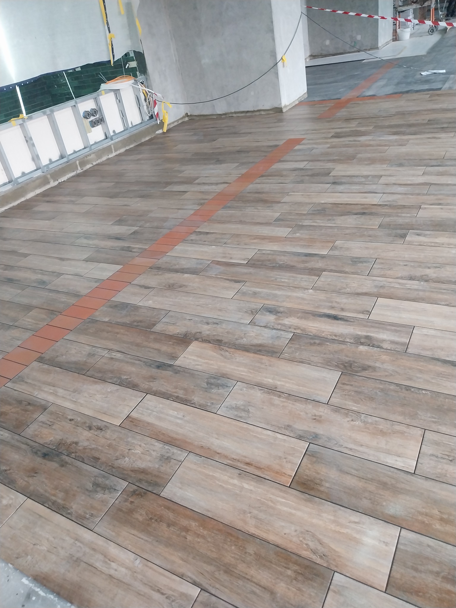 Ułożona podłoga z płytek imitujących drewno, z pomarańczowym pasem płytek oddzielającym strefy, widoczne prace wykończeniowe ścian w tle.