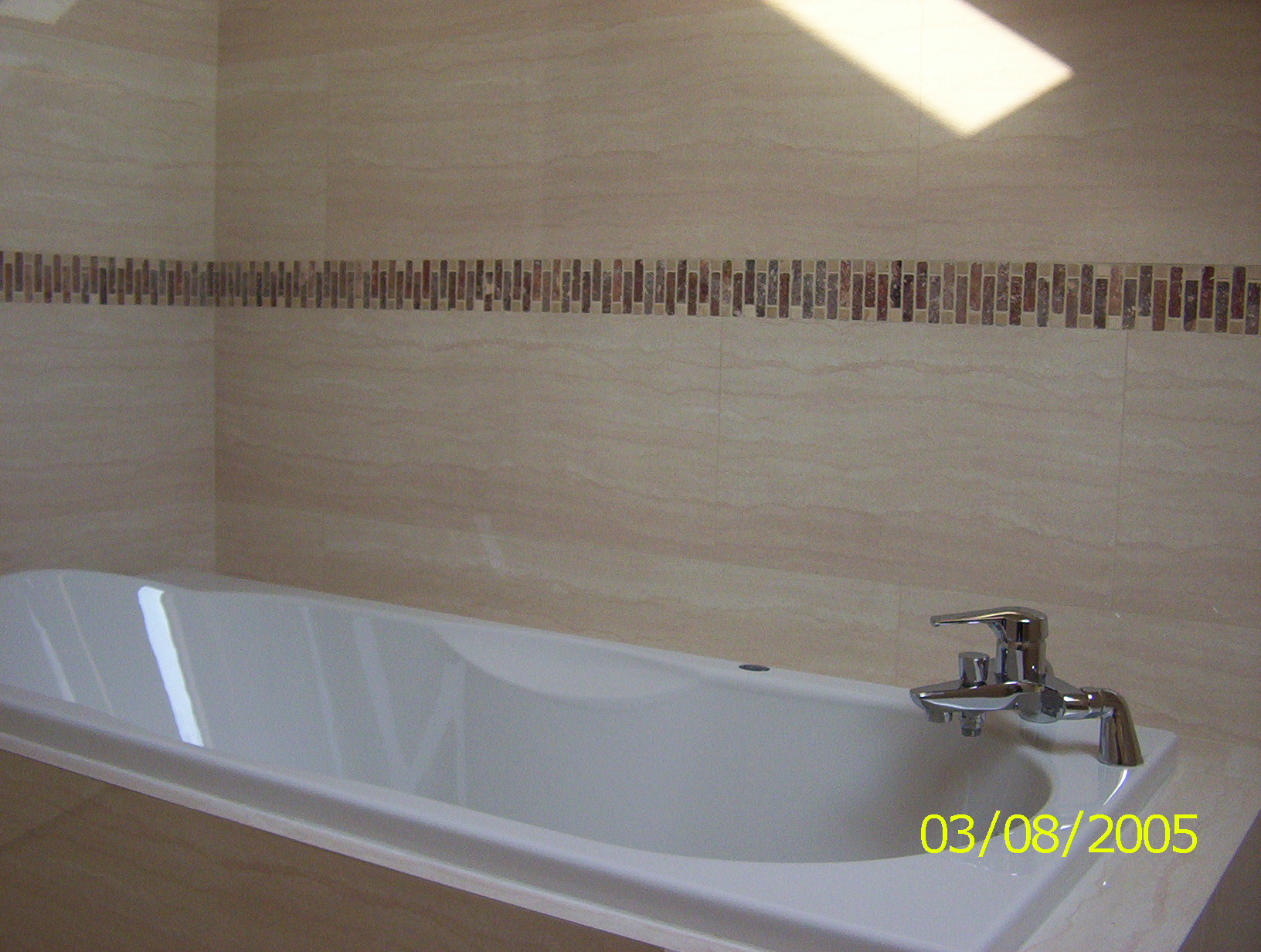 Wykończona łazienka z wanną i beżowymi płytkami z dekoracyjnym pasem mozaiki z brązowych i beżowych płytek.