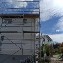 Elewacja domu jednorodzinnego w trakcie prac wykończeniowych, osłonięta rusztowaniem z siatką zabezpieczającą, widok z poziomu ogrodu.