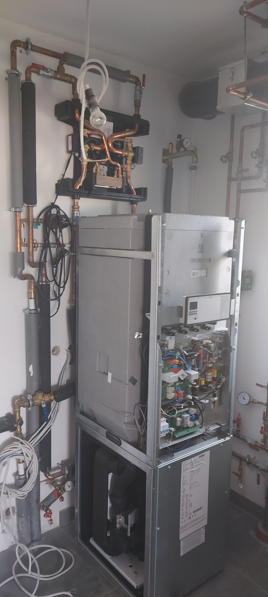 Nowoczesny piec CO z otwartą obudową, widoczne wewnętrzne komponenty i podłączenia rur miedzianych, z tymczasowym oświetleniem żarówką.