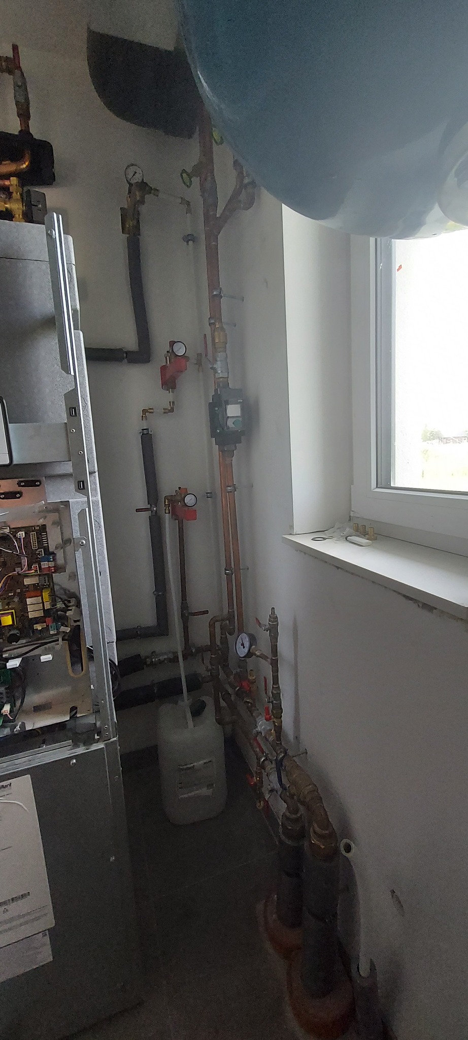 Wnętrze kotłowni z otwartym kotłem gazowym, instalacją hydrauliczną z rur miedzianych, zbiornikiem wyrównawczym i osprzętem pomiarowym przy ścianie z oknem.