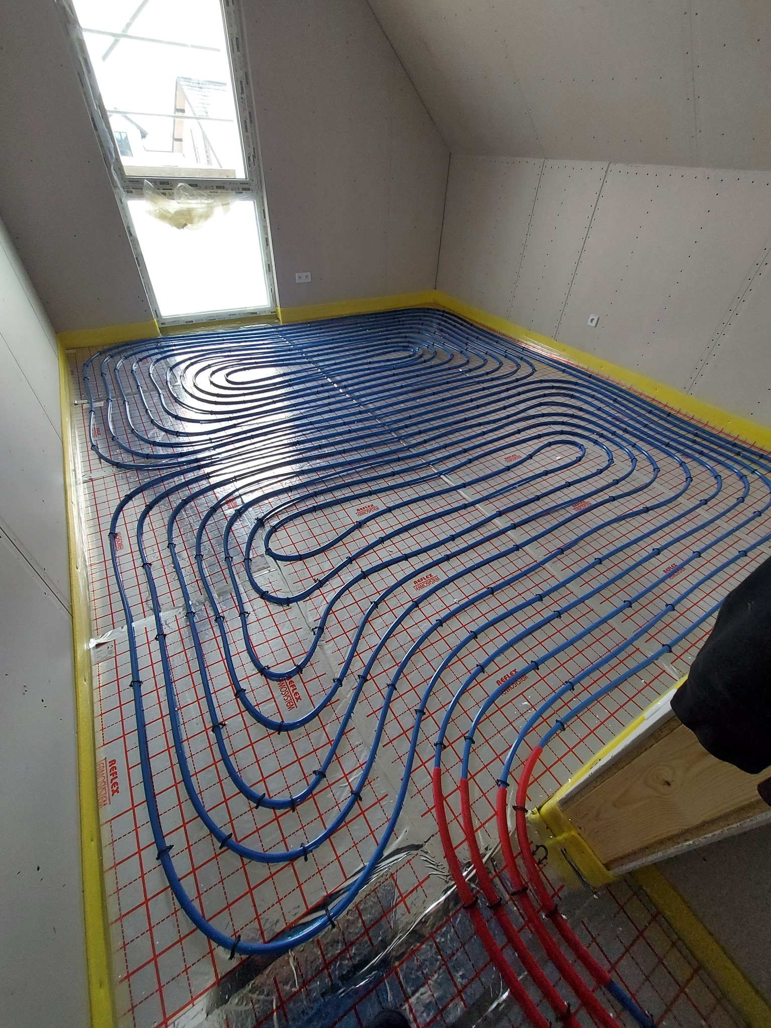 Niebieskie rury ogrzewania podłogowego ułożone na folii refleksyjnej z czerwonym nadrukiem siatki, widok z góry w pomieszczeniu na poddaszu z oknem dachowym.